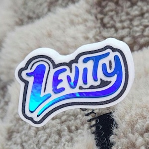 Può includere: Un badge bianco con la scritta "Levity" in lettere blu e nere. Il badge ha una clip a molla nera ed è attaccato a una superficie sfocata di colore chiaro. La parola "Levity" è in un carattere stilizzato.