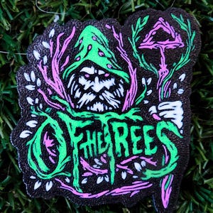 Può includere: Toppa nera con la scritta "OF THE REES" in verde, raffigurante una figura con cappello verde e barba bianca, circondata da fogliame rosa e bianco e un fungo. La toppa è su una superficie erbosa verde.