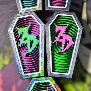 Può includere: Una collezione di spille a forma di bara con un design a strati. Ogni spilla presenta un emblema astratto stilizzato in colori vivaci come rosa, verde e blu. Le spille hanno una finitura metallica iridescente e un bordo bianco.
