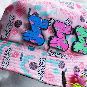 May include: Pink bucket hat with a repeating pattern of shapes and palm fronds. The hat features three colorful dinosaur-shaped patches in blue, pink, and green. The hat is displayed on a white mannequin head.