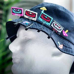 May include: A dark blue bucket hat adorned with several cassette tape-shaped pins in pink, white, and blue. Each pin features a monster face design and the text "TAPE 8". Additional mushroom-shaped pins are also attached.