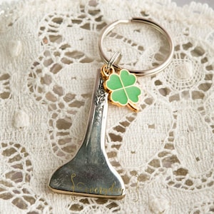 May include: A silver fork-shaped keyring with a gold-coloured rim and a green four-leaf clover charm. The keyring has a silver ring and is set against a white lace background. The word "everyday" is printed on the fork.