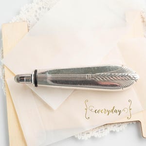 May include: A silver letter opener with an arrow and wheat design, resting on a stack of cream-colored paper and a wooden base. The word "everyday" is written in gold script on the paper.