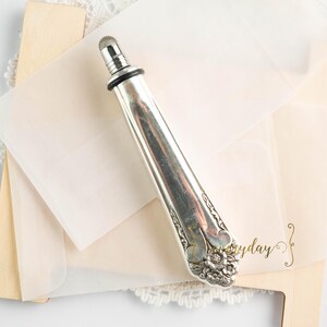 May include: A silver bottle stopper with a decorative floral design, resting on a stack of cream-colored paper and a wooden element. The stopper is approximately 6 inches long.