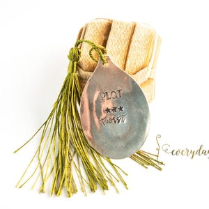 Peut inclure: Un pendentif argenté en forme de goutte d'eau, estampillé des mots "PLOT" et "TWIST" et de trois étoiles. Il est suspendu à des glands verts et repose sur un objet beige texturé. Le mot "everyday" est écrit en cursive.