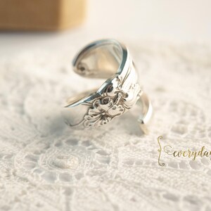 May include: A silver spoon ring with floral detailing. The ring is crafted from a spoon, with the handle forming the band and the bowl of the spoon creating a decorative element. The ring is displayed on a white lace surface.