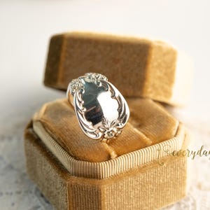 May include: A silver ring with an oval, ornate design, resting on a tan velvet ring box. The ring features detailed floral and scrollwork patterns. The background includes a second ring box and a lace-like fabric.