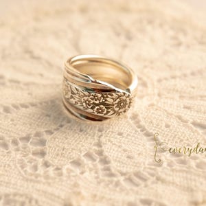 May include: A silver ring with a floral design. The ring is made from a spoon and has a detailed floral pattern around the band. The ring is set on a white lace surface.