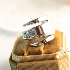 May include: A handcrafted silver spoon ring with a floral design. The ring is wrapped in a spiral shape and rests on a gold velvet ring box. The ring is a unique piece of jewelry.
