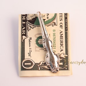 May include: A silver fork money clip holding a one-dollar bill. The bill is green and tan, with the words "United States of America" and the number "1" visible. The fork has an ornate design.