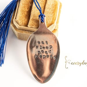 May include: A spoon-shaped bookmark with the words "eat sleep read repeat" stamped on it. The bookmark has a blue tassel and is next to a gold-colored box. The word "everyday" is written in cursive.