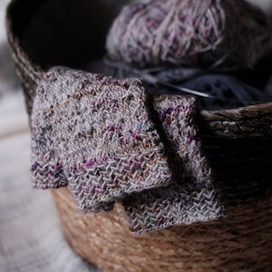 May include: A woven basket filled with a ball of yarn and two pairs of knitted fingerless gloves. The gloves are a speckled grey and pink color.