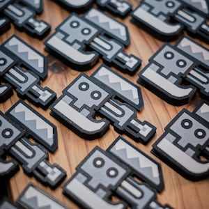 May include: A close-up of multiple gray and white 3D printed robot figurines with a pixelated design. The figurines are arranged on a wooden surface.