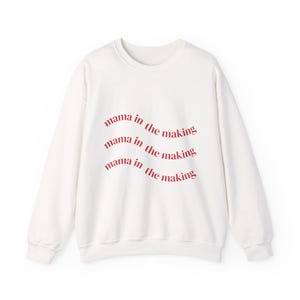 May include: White crew neck sweatshirt with the phrase "mama in the making" in red script across the front. The text is repeated three times in a curved, wave-like pattern. The sweatshirt has long sleeves and a relaxed fit.
