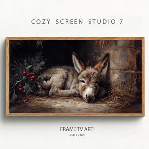 Puede incluir: Una impresión artística digital enmarcada que representa un burro descansando en un establo. La obra de arte tiene una estética rústica, con el burro en tonos grises y marrones, y una ramita de acebo con bayas rojas. El texto "COZY SCREEN STUDIO 7" y "FRAME TV ART" son visibles.