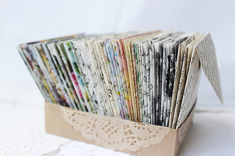 May include: A collection of small, handmade envelopes with various patterned paper designs, arranged in a brown box with a lace trim.