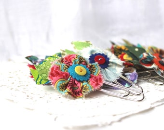May include: A set of six colorful fabric-covered paper clips with buttons. The clips are decorated with floral patterns and various colors, including pink, blue, green, and yellow.