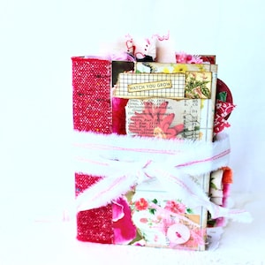 May include: A handmade journal with a red textured fabric spine and a cover featuring floral patterns, vintage text, and a label with the text WATCH YOU GROW. The book is tied with a white frayed fabric ribbon and includes layered paper elements.