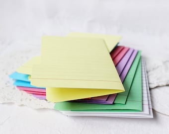 May include: A stack of colorful, lined index cards in shades of yellow, pink, blue, green, and purple. The cards are fanned out, revealing their edges and the red lines. The background is a soft, white surface.