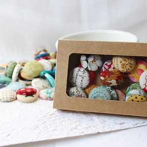 May include: A brown cardboard box with a clear window displaying an assortment of colourful fabric-covered buttons. The buttons feature various patterns and designs, including floral, geometric, and abstract.