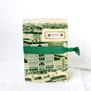 Peut inclure: Un carnet de notes de style vintage avec une illustration en noir et blanc d'une scène de ville. Il est fermé par un élastique vert. Le mot "NOTES" est imprimé sur une étiquette.