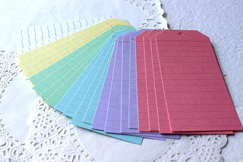 May include: A set of colorful lined paper gift tags. The tags are arranged in a fan shape, with the colors ranging from white to pink. The tags are all lined with a thin black line.