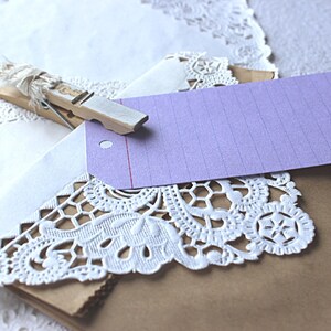 May include: A brown paper gift bag with a white doily and a purple gift tag attached with a wooden clothespin. The tag has a red line and lined paper.