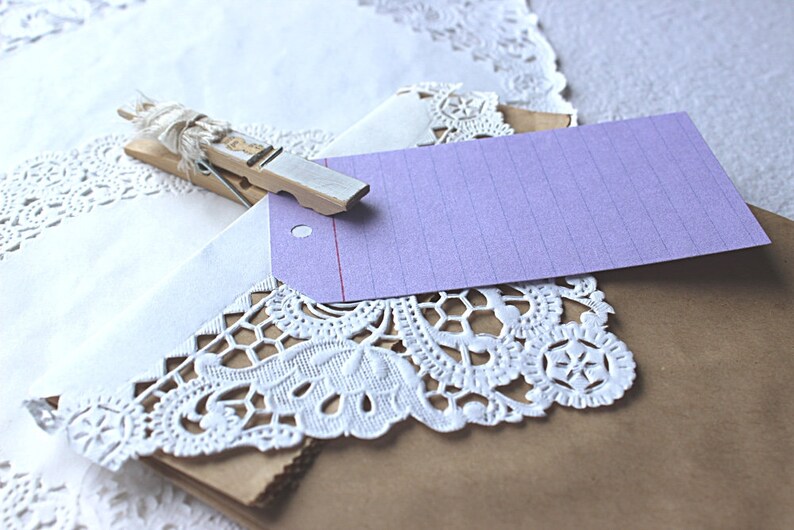 May include: A brown paper gift bag with a white doily and a purple gift tag attached with a wooden clothespin. The tag has a red line and lined paper.