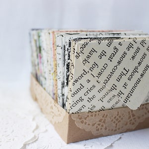 May include: A small cardboard box with a lace trim, filled with stacks of paper cards. The cards are made from recycled book pages and feature text from the book. The text on the top card reads: "Then the snow-covered mountain's snow-shoes at his war-old snow."