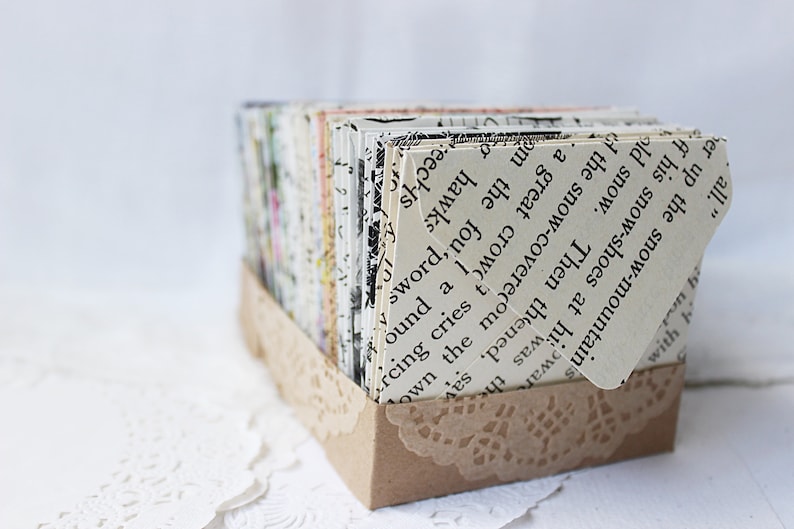 May include: A small cardboard box with a lace trim, filled with stacks of paper cards. The cards are made from recycled book pages and feature text from the book. The text on the top card reads: "Then the snow-covered mountain's snow-shoes at his war-old snow."