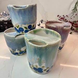 May include: Four ceramic mugs in shades of blue, green, and purple. Each mug has a unique glaze and a handle-like opening at the top. The bases are decorated with blue floral patterns and green leaves. The mugs are arranged on a white surface.