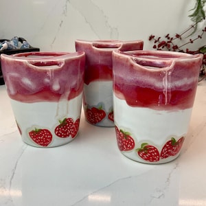 May include: Three ceramic mugs with a white base and a red and pink ombre effect at the top. The mugs have a handle cutout and are decorated with strawberry decals around the base. The mugs are on a white surface.