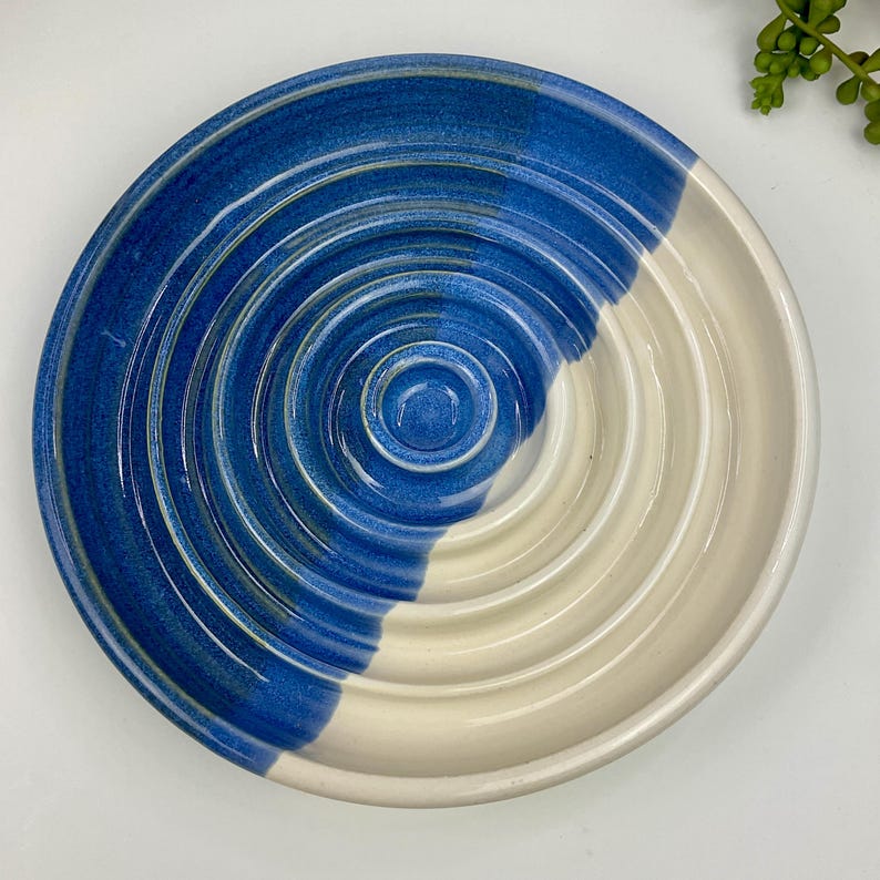 May include: A round ceramic dish featuring a concentric ribbed pattern. The surface is divided into two distinct colour sections, with one half glazed in a deep cobalt blue and the other half in a smooth, off-white cream finish.