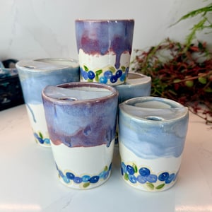Op de afbeelding: Een verzameling van vijf keramische bekers met deksels, voorzien van een witte basis met handgeschilderde blauwe bessen aan de onderkant. De bovendelen zijn geglazuurd in diverse tinten blauw en paars, met een kenmerkende druipende afwerking.