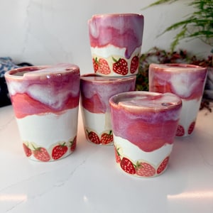 May include: A collection of five ceramic cups featuring a white base with a hand-painted strawberry pattern around the bottom and a glossy, drippy pink and white glaze finish on the upper portion. Each cup has a small notch on the rim.