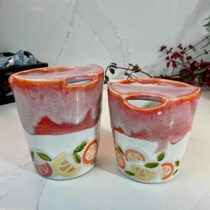 Puede incluir: Dos tazas de cerámica con un diseño único. La mitad superior de cada taza presenta un degradado de rojo y blanco, mientras que la mitad inferior es blanca con un patrón de frutas cítricas de colores. Cada taza tiene una abertura para el asa en la parte superior.