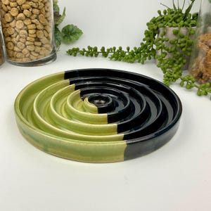 May include: A circular ceramic slow feeder bowl featuring a concentric ring design. The bowl is finished in a two-tone glaze, with one half in a vibrant lime green and the other half in a deep, glossy black, creating a distinct colour split.