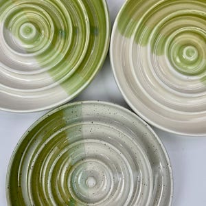 May include: Three circular ceramic plates featuring concentric ring patterns. Two plates display a smooth cream and light green gradient, while the third plate features a cream base with green speckles and a solid green band along one side.