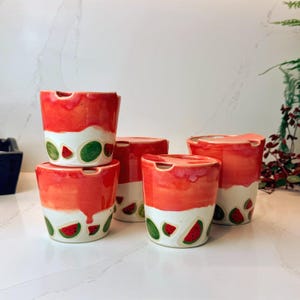 Pode incluir: Um conjunto de cinco canecas de cerâmica com um design de melancia. As canecas são vermelhas e brancas com detalhes de fatias de melancia verdes e vermelhas. Cada caneca tem uma pequena abertura curva no topo.
