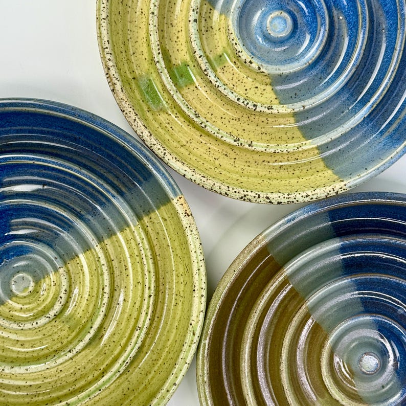 May include: Three circular ceramic bowls featuring a concentric ridged texture. The surfaces are finished with a speckled glaze in shades of deep blue, bright yellow, and earthy brown, creating a distinct colour-blocked effect across each bowl.