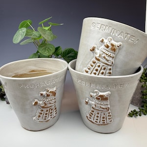 Puede incluir: Un juego de cuatro macetas de cerámica blanquecina. Cada maceta presenta una imagen en relieve marrón de un Dalek y una palabra: "PROPAGATE!", "POLLINATE!" y "GERMINATE!". Las macetas son de diferentes alturas y formas, con una estética rústica.