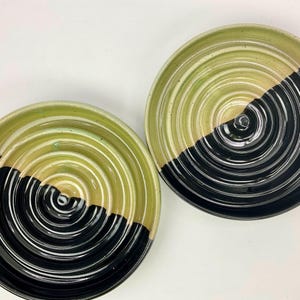 May include: Two round ceramic dishes featuring a concentric circular grooved pattern. Each dish is finished with a two-tone glaze, displaying a vibrant lime green on one half and a glossy solid black on the other half, creating a distinct contrast.