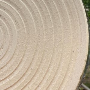 May include: A circular ceramic plate with a light beige, matte finish featuring concentric, grooved rings radiating from the centre. The surface has a textured, granular appearance with visible clay particles throughout the material.