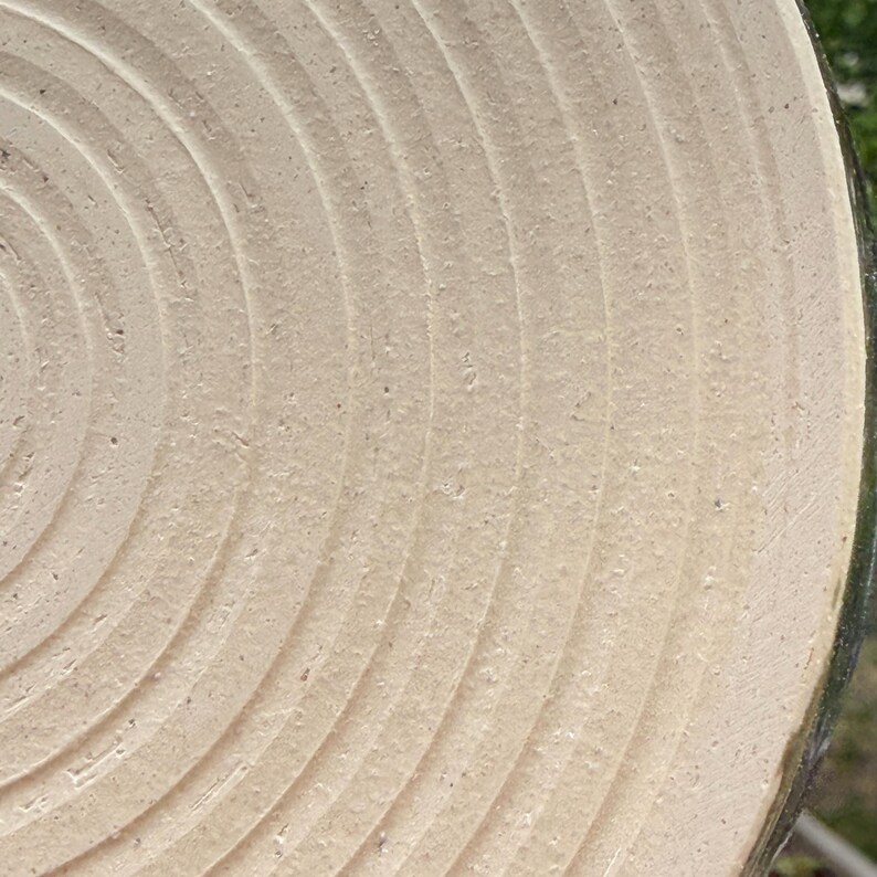 May include: A circular ceramic plate with a light beige, matte finish featuring concentric, grooved rings radiating from the centre. The surface has a textured, granular appearance with visible clay particles throughout the material.