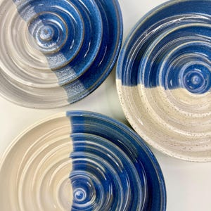 May include: Three circular ceramic bowls featuring a concentric ridged design. Each bowl is finished with a two-tone glaze, split between a speckled off-white and a deep blue. The surfaces exhibit a glossy texture with visible circular grooves.