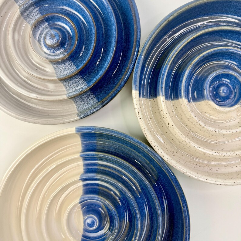 May include: Three circular ceramic bowls featuring a concentric ridged design. Each bowl is finished with a two-tone glaze, split between a speckled off-white and a deep blue. The surfaces exhibit a glossy texture with visible circular grooves.