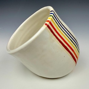 May include: White ceramic bowl with a unique, asymmetrical shape. A series of colorful stripes in red, orange, yellow, green, blue, and purple adorn the side. The bowl has a smooth, glossy finish and a minimalist design.