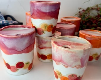 May include: A collection of ceramic cups featuring a white base with a painted pattern of red, orange, and yellow tomatoes near the bottom. Each cup has a pinkish-red glazed upper section and a semi-circular lid with a small circular opening.