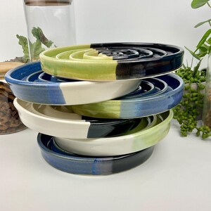 May include: A stack of five circular ceramic soap dishes with raised spiral ridges. Each dish features a two-tone glazed finish in combinations of lime green, deep blue, and off-white, with smooth, rounded edges and a glossy, reflective surface.