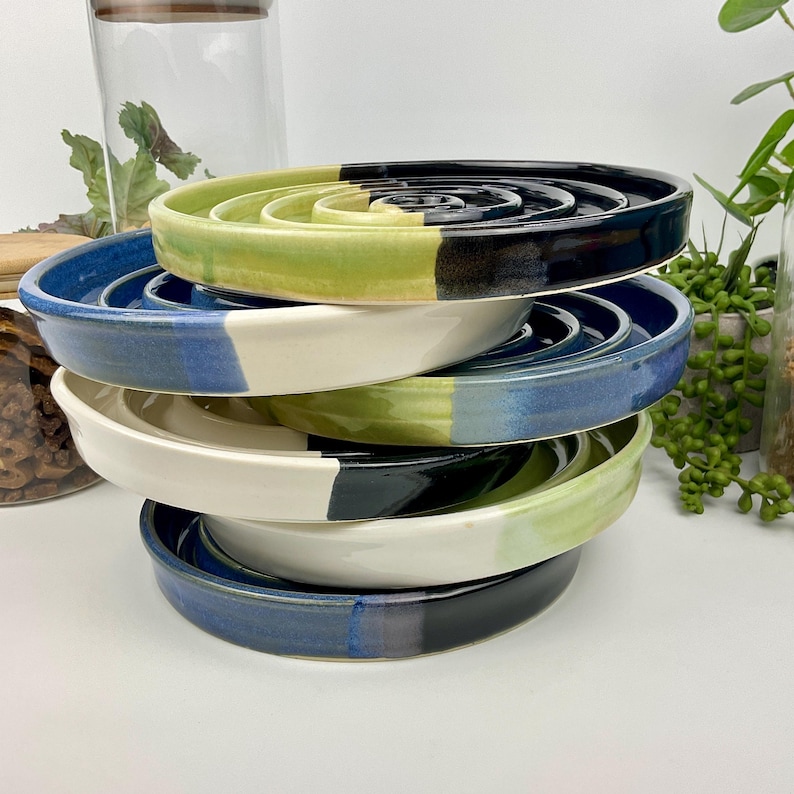 May include: A stack of five circular ceramic soap dishes with raised spiral ridges. Each dish features a two-tone glazed finish in combinations of lime green, deep blue, and off-white, with smooth, rounded edges and a glossy, reflective surface.